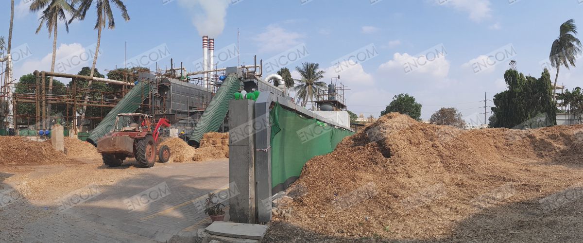Biomass boiler site with operating boiler and large piles of biomass fuel under clear sky